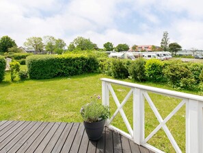 A wooden deck with a railing, a potted plant, and a view of a grassy area with a hedge and parked vehicles in the background.