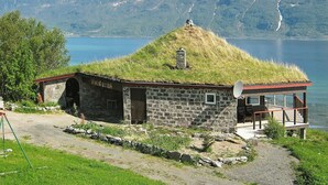 A stone building with a grass roof, a satellite dish, and a view of a lake and mountains in the background. - 6 Person Holiday Home in Hamnvik (Ibestad)