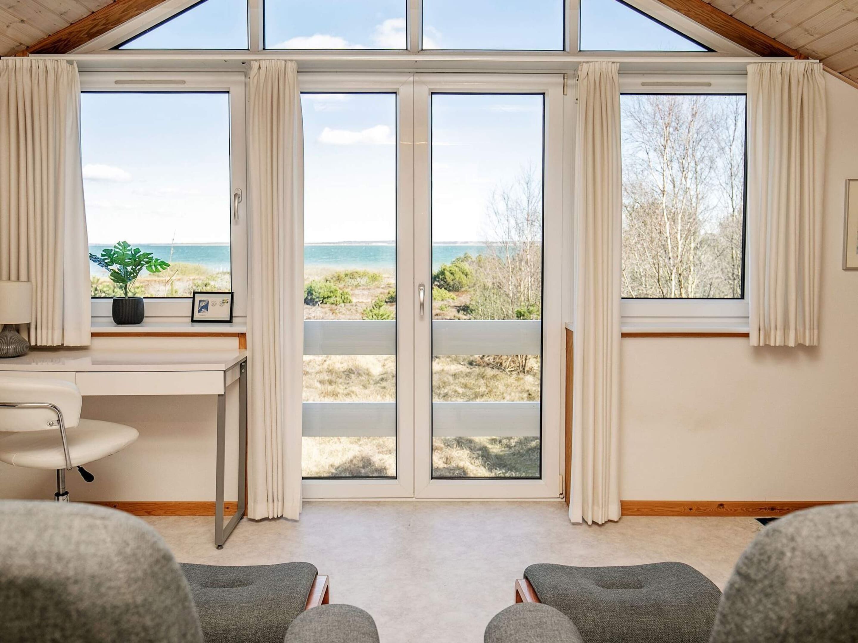Une pièce dotée d’une grande porte-fenêtre coulissante donnant sur la plage, d’un bureau avec une chaise et de deux fauteuils gris.