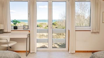 Une pièce dotée d’une grande porte-fenêtre coulissante donnant sur la plage, d’un bureau avec une chaise et de deux fauteuils gris.