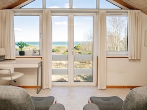 A room with a large sliding glass door leading to a beach view, a desk with a chair, and two grey armchairs. - 4 Star Holiday Home in Thyholm (Thyholm)