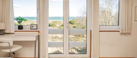 A room with a large sliding glass door leading to a beach view, a desk with a lamp, and two grey armchairs.