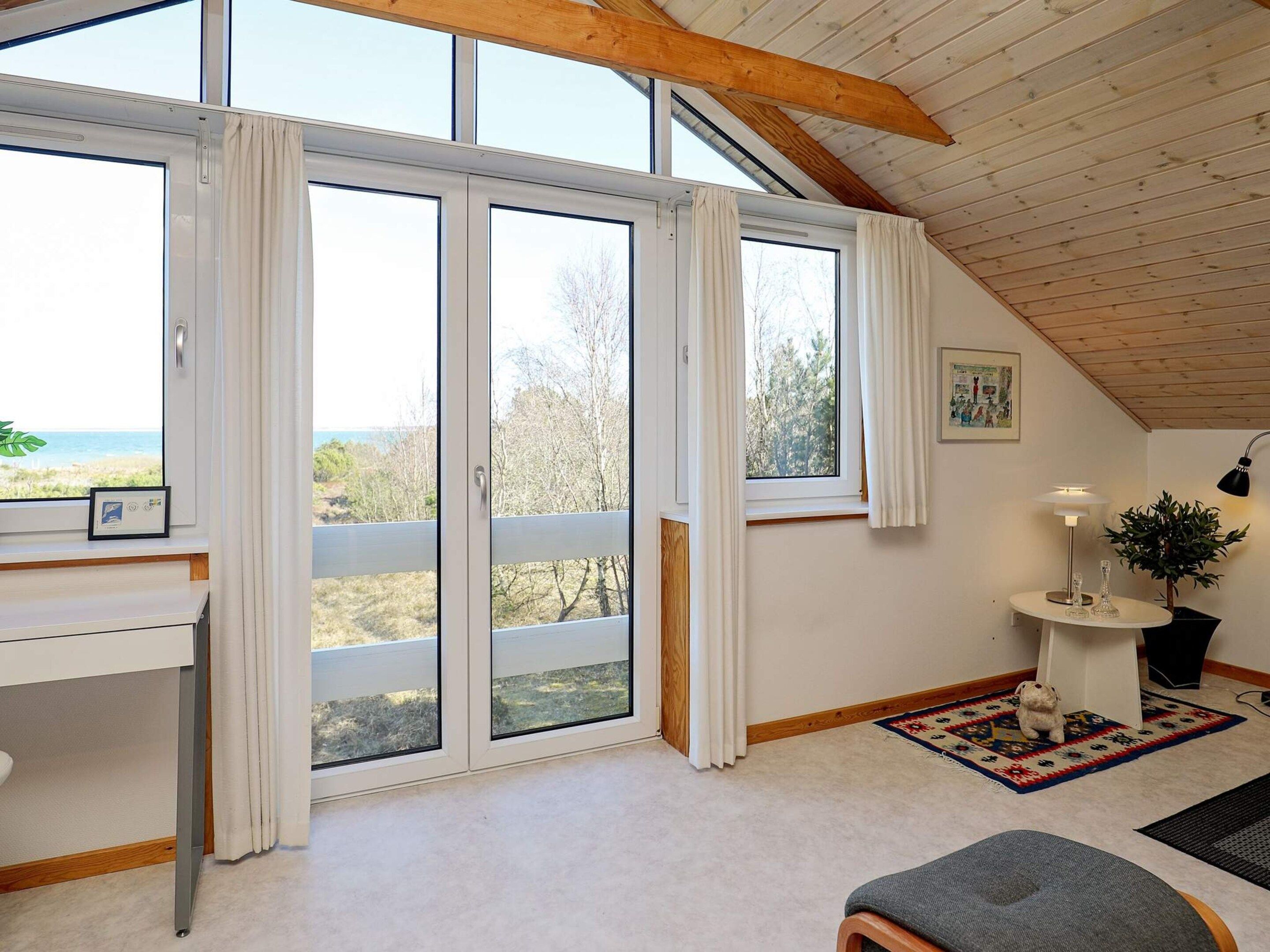 A room with large windows, a desk, a small table with a lamp, and a rug.