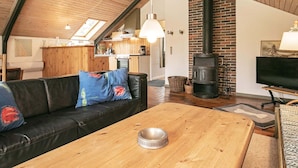 Ein gemütliches Wohnzimmer mit einer schwarzen Ledersofa, einem Holztisch und einem Fernseher. Im Hintergrund sind ein Kamin aus Ziegelsteinen und ein Oberlicht zu sehen.