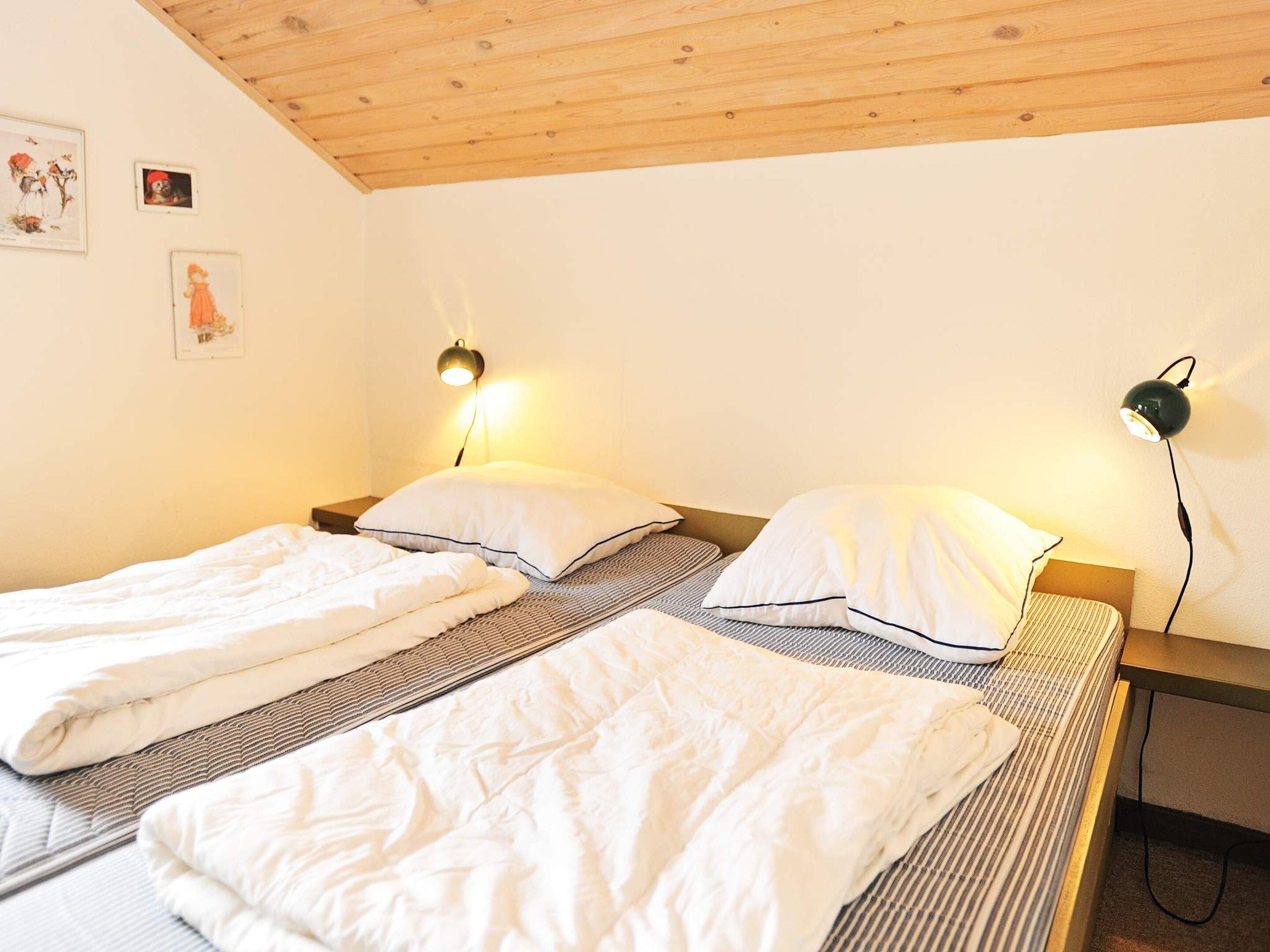 A room with two single beds, each with a pillow and a blanket, a wooden ceiling, and wall-mounted lamps.