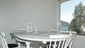 A dining area with a round table and four white chairs, a patterned tablecloth, and a view of a body of water through a window. - Holiday Home in Åfarnes (Rauma)