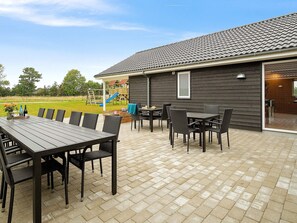 An outdoor patio area with a table and chairs, a wooden play structure in the background, and a building with a tiled roof. - Spacious Holiday Home in Grenå near Sea (Grenaa)