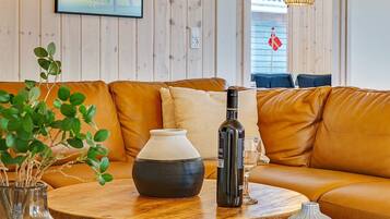 Una sala de estar acogedora con un sofá de cuero marrón, una mesa de centro redonda de madera, una botella de vino, una copa y un florero con plantas verdes.