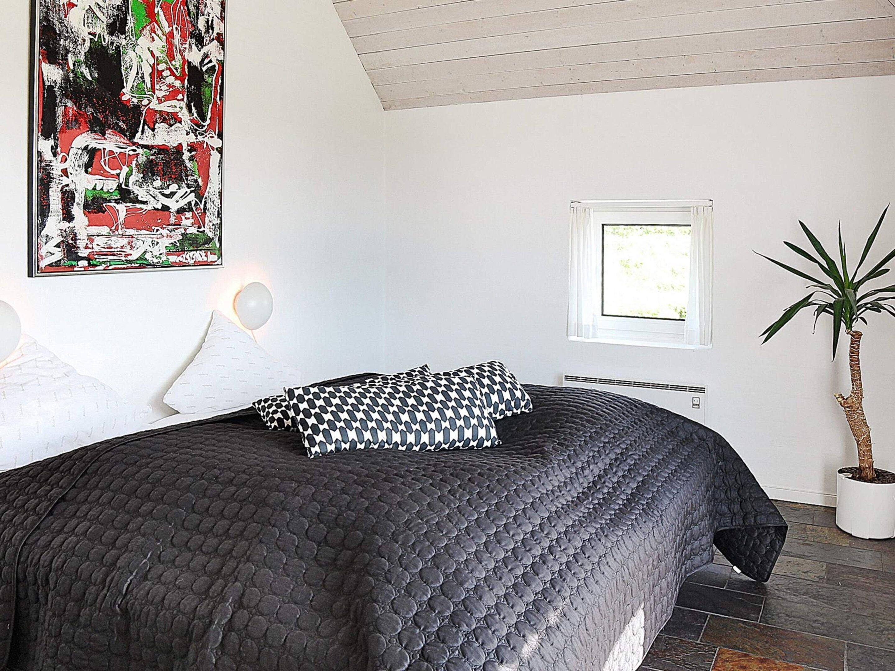 A bedroom with a large bed, a potted plant, a window, and a wall-mounted artwork.