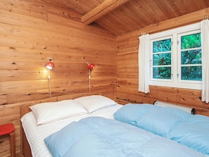 A wooden cabin room with a bed, two bedside lamps, a window with curtains, and a view of greenery outside.