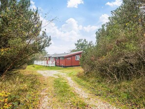 A red cabin with a gravel path leading to it, surrounded by trees and shrubs. - 6 Person Holiday Home in Oksbol (Oksbol)