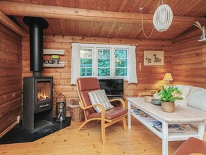 A cozy cabin interior with a wood-burning stove, a comfortable chair, a sofa, a table, and a window with a view of greenery. - 6 Person Holiday Home in Oksbol (Oksbol)