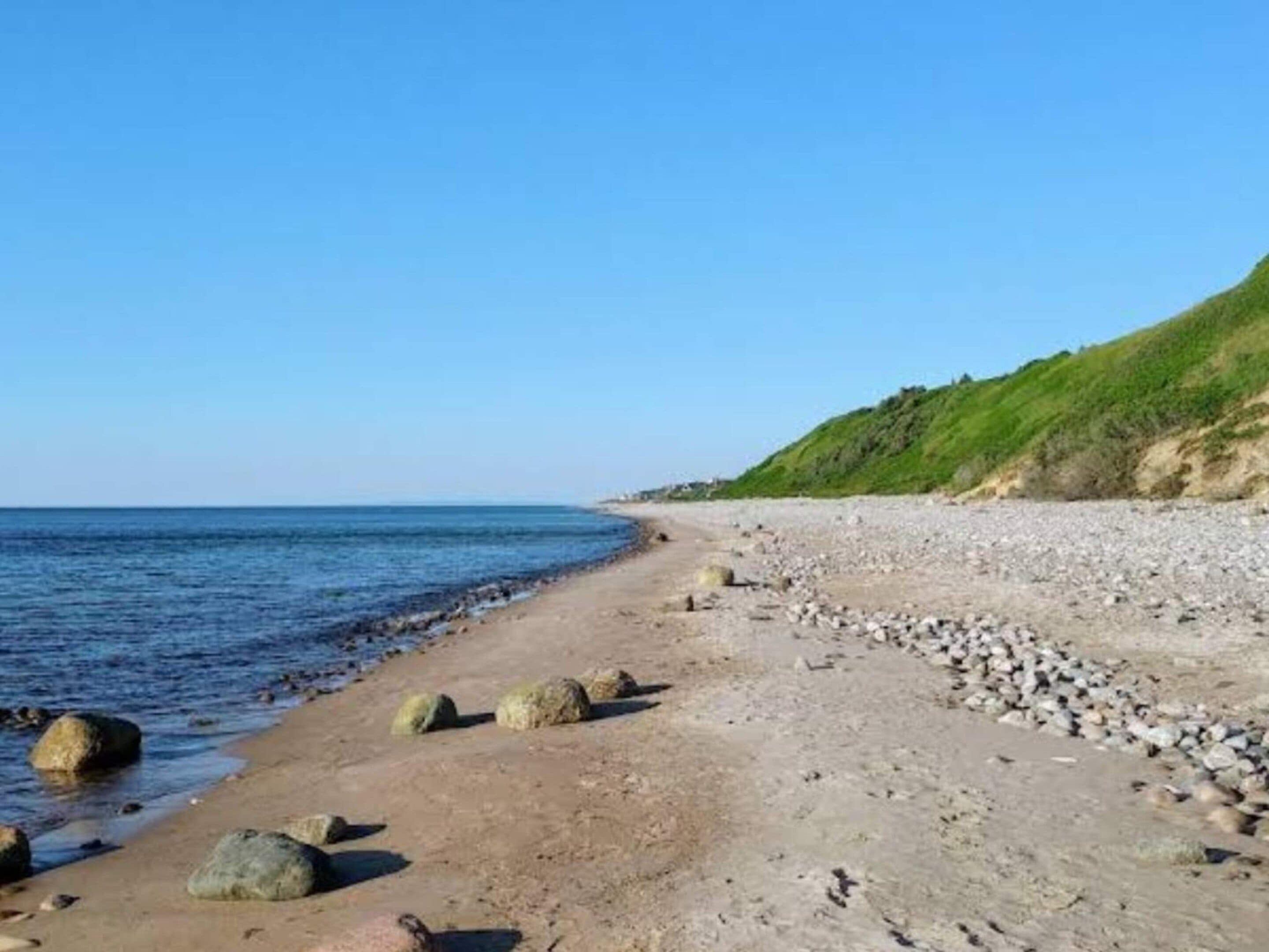 A beach with a clear sky, a sandy shore with scattered rocks, and a grassy hill in the background.