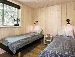 Una habitación con dos camas, paredes de madera, una ventana con plantas y una mesita de noche pequeña.