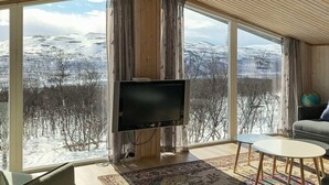 A room with a large window offering a snowy mountain view, a flat-screen TV, a small round table, and a sofa. - Holiday Home in Kvalsund (Hammerfest)