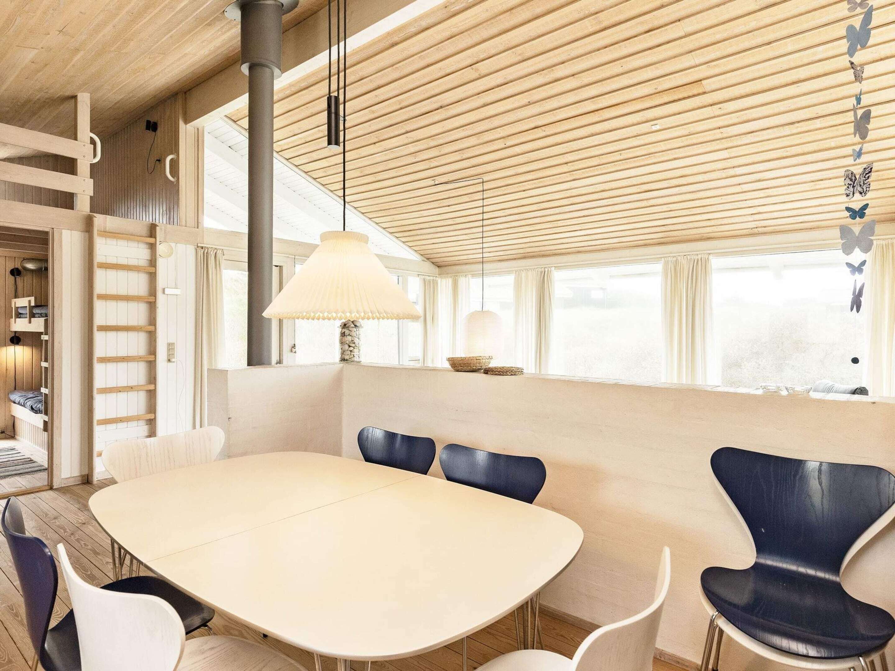 Uma sala de jantar moderna com uma grande mesa oval, cadeiras azuis e brancas, uma luminária pendente e vista para o exterior através de amplas janelas.