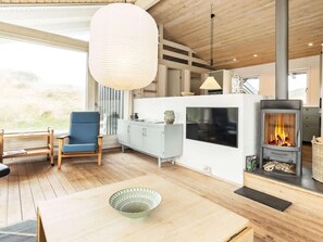 A modern living room with a wooden floor, a blue chair, a white cabinet, a large hanging light fixture, and a wood-burning stove. - 4 Star Holiday Home in Pandrup (Pandrup)