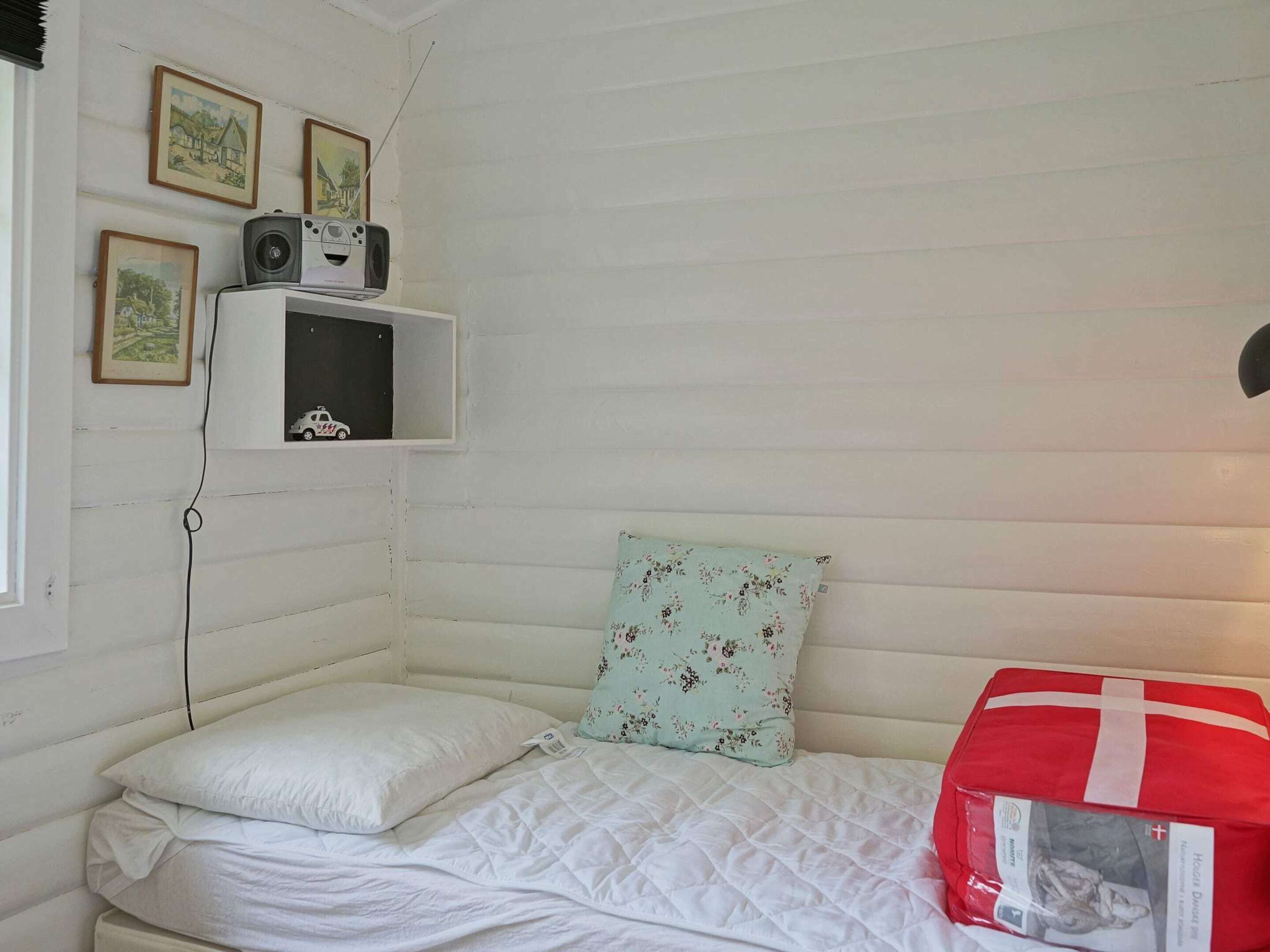 Una habitación pequeña con una cama individual, cabecera blanca, colcha acolchada, almohada estampada con motivos florales, una pequeña estantería con fotografías enmarcadas y una caja roja a la derecha.