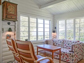 A cozy room with a wooden chair, a floral sofa, and a table. There are large windows with a view of trees. - 4 Person Holiday Home in Allinge (Allinge)
