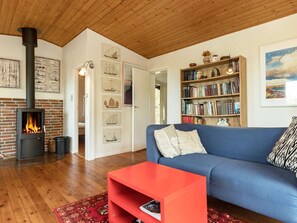 A cozy living room with a brick fireplace, a blue sofa, a red coffee table, and a bookshelf filled with books. - 4 Person Holiday Home in Martofte-by Traum (Martofte)