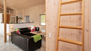 Interior acolhedor de uma cabana, com teto de madeira, sofá de couro preto, mesa de jantar de madeira e área de cozinha visível através de uma porta aberta.