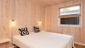 Quarto com parede revestida de madeira, cama com roupa de cama branca, duas almofadas padronadas em preto e branco, mesa de cabeceira e janela com persianas.