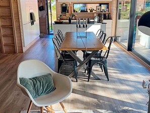 A modern dining area with a wooden table and chairs, a white armchair with a green cushion, and a kitchen in the background.