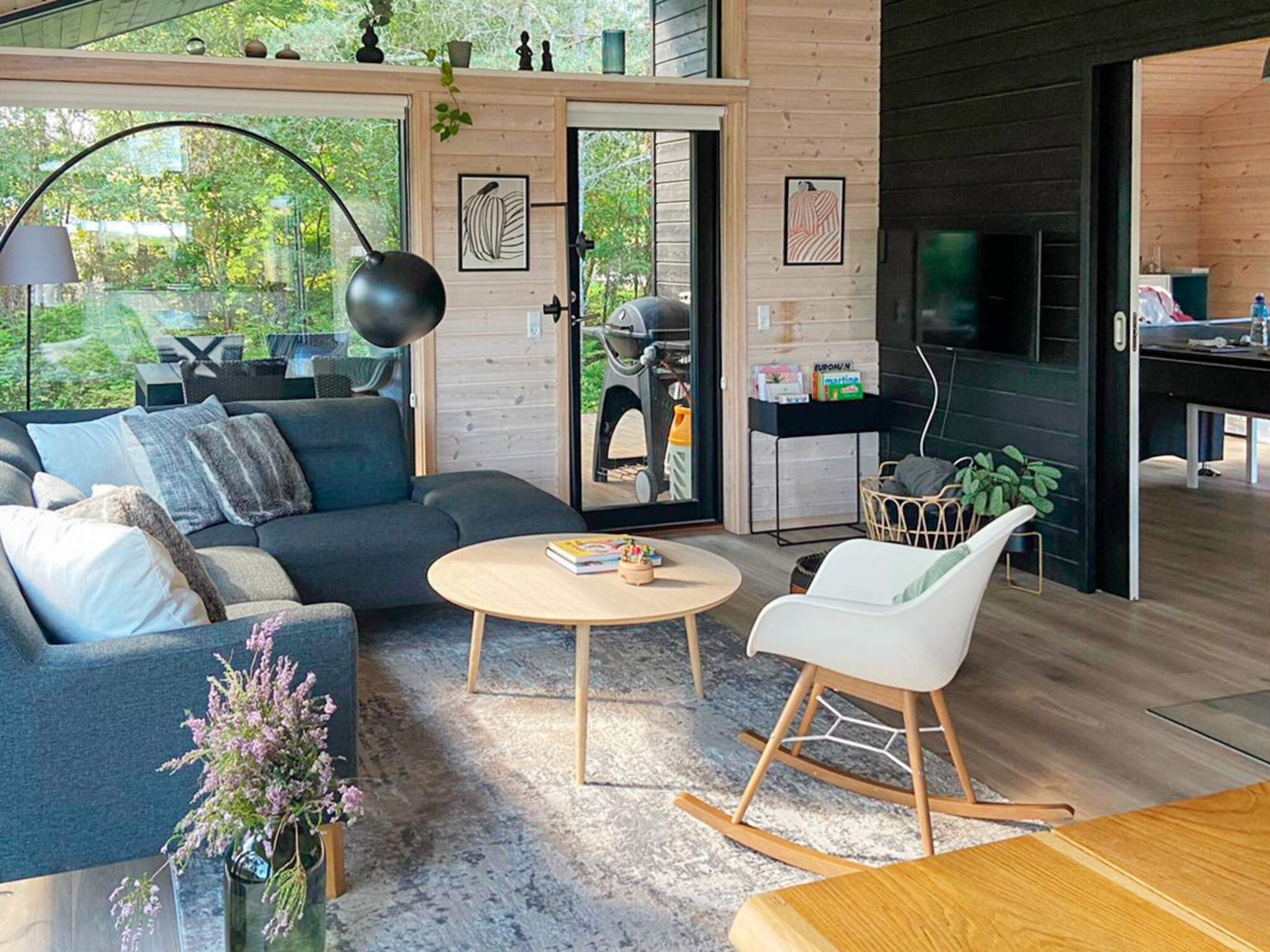 A modern living room with a sofa, a round coffee table, and a rocking chair. There is a door leading to a balcony with a view of greenery.