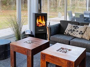 A cozy outdoor seating area with a wood-burning stove, two wooden tables with decorative inlays, a cushioned sofa, and a view of the surrounding nature.