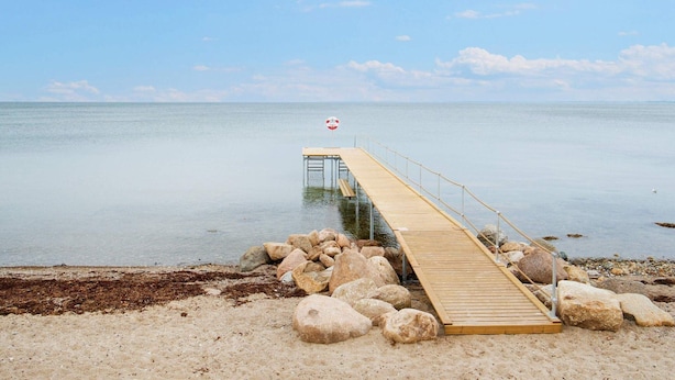 Eine Holzstege ragt in ruhiges Wasser, umgeben von Felsen und Kieselsteinen.