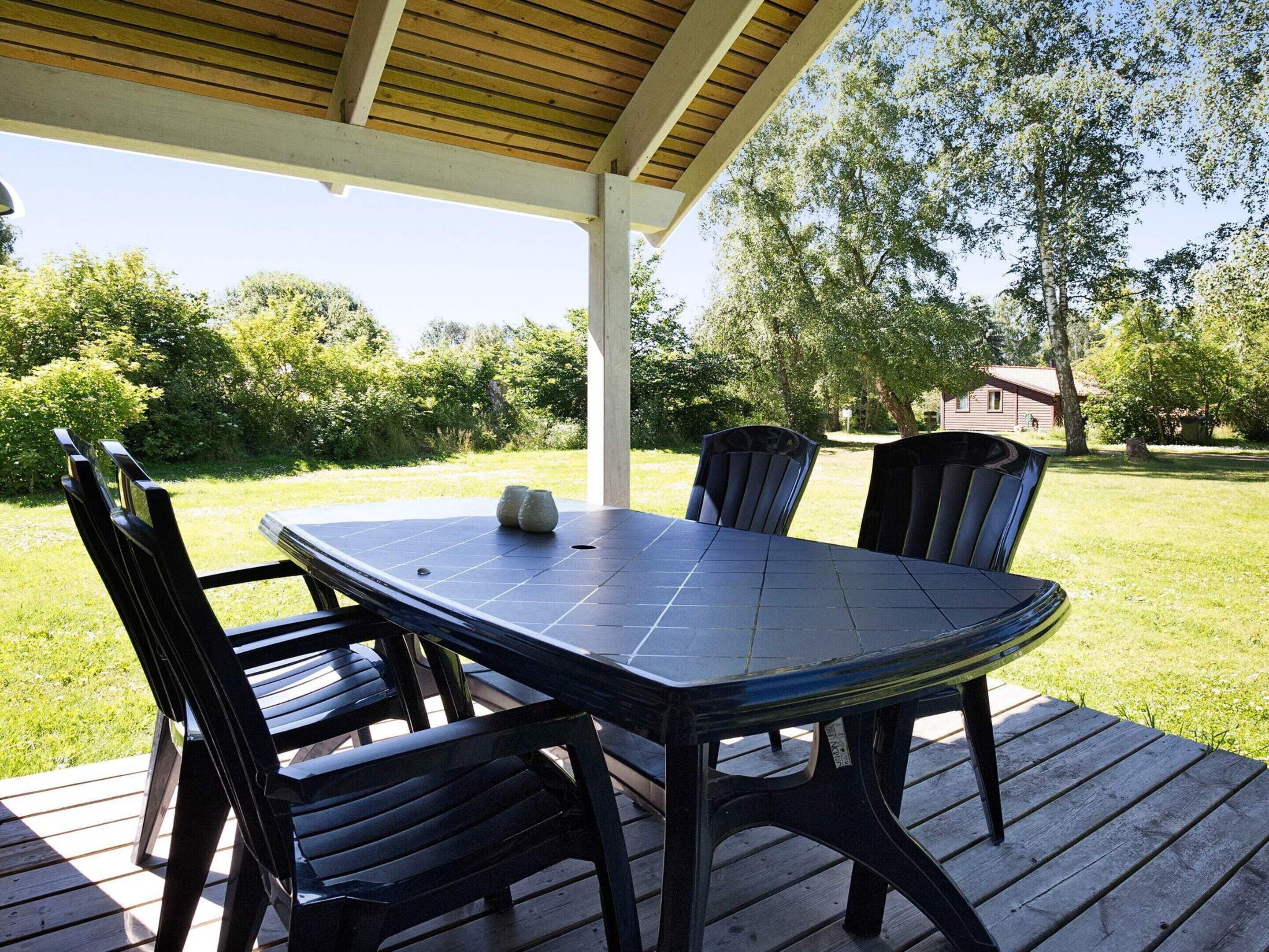 Ein überdachter Essbereich im Freien mit Tisch und Stühlen, umgeben von einer Holzterrasse und Grünpflanzen.