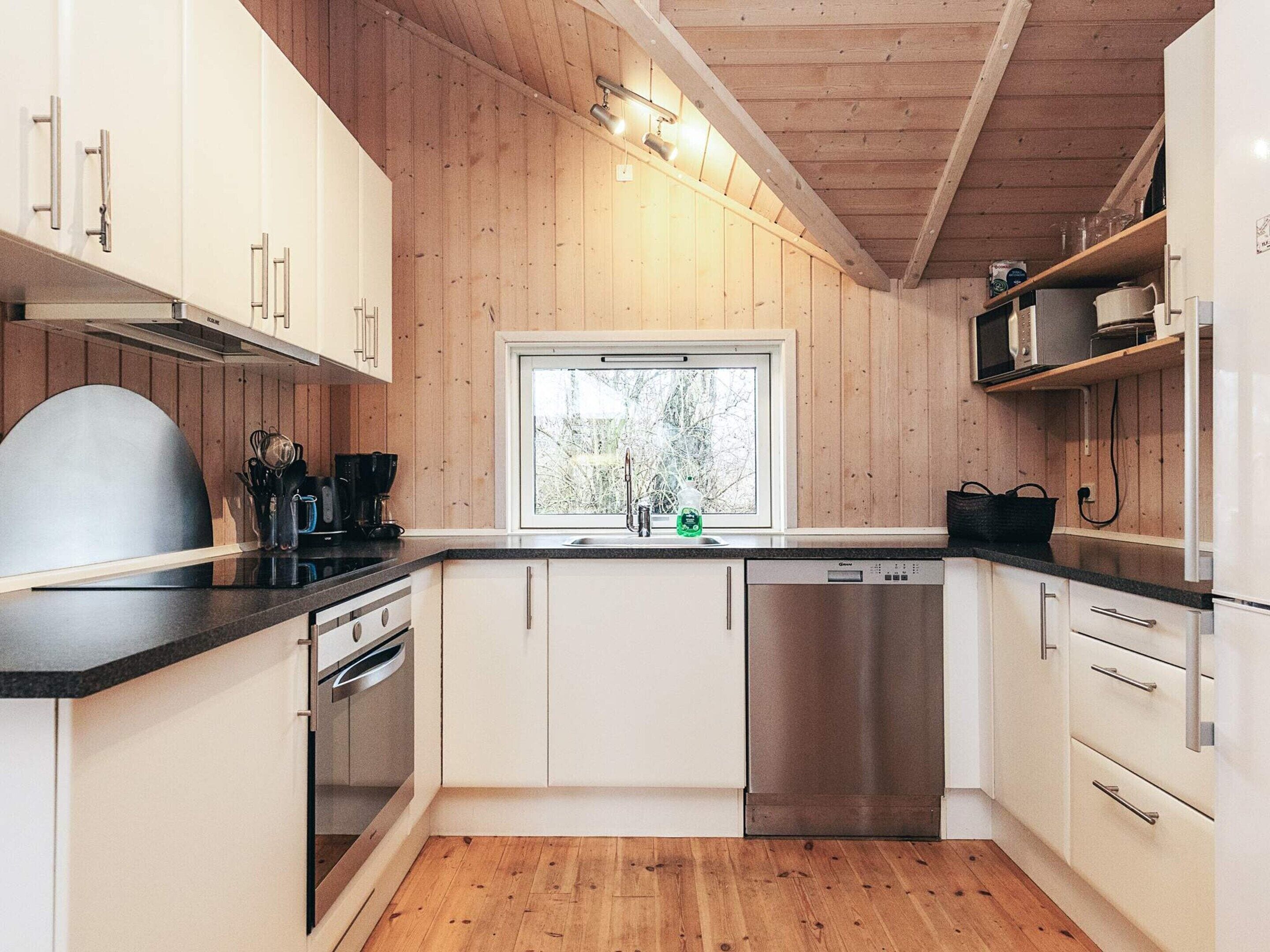 A modern kitchen with wooden walls, a window, stainless steel appliances, and a black countertop.
