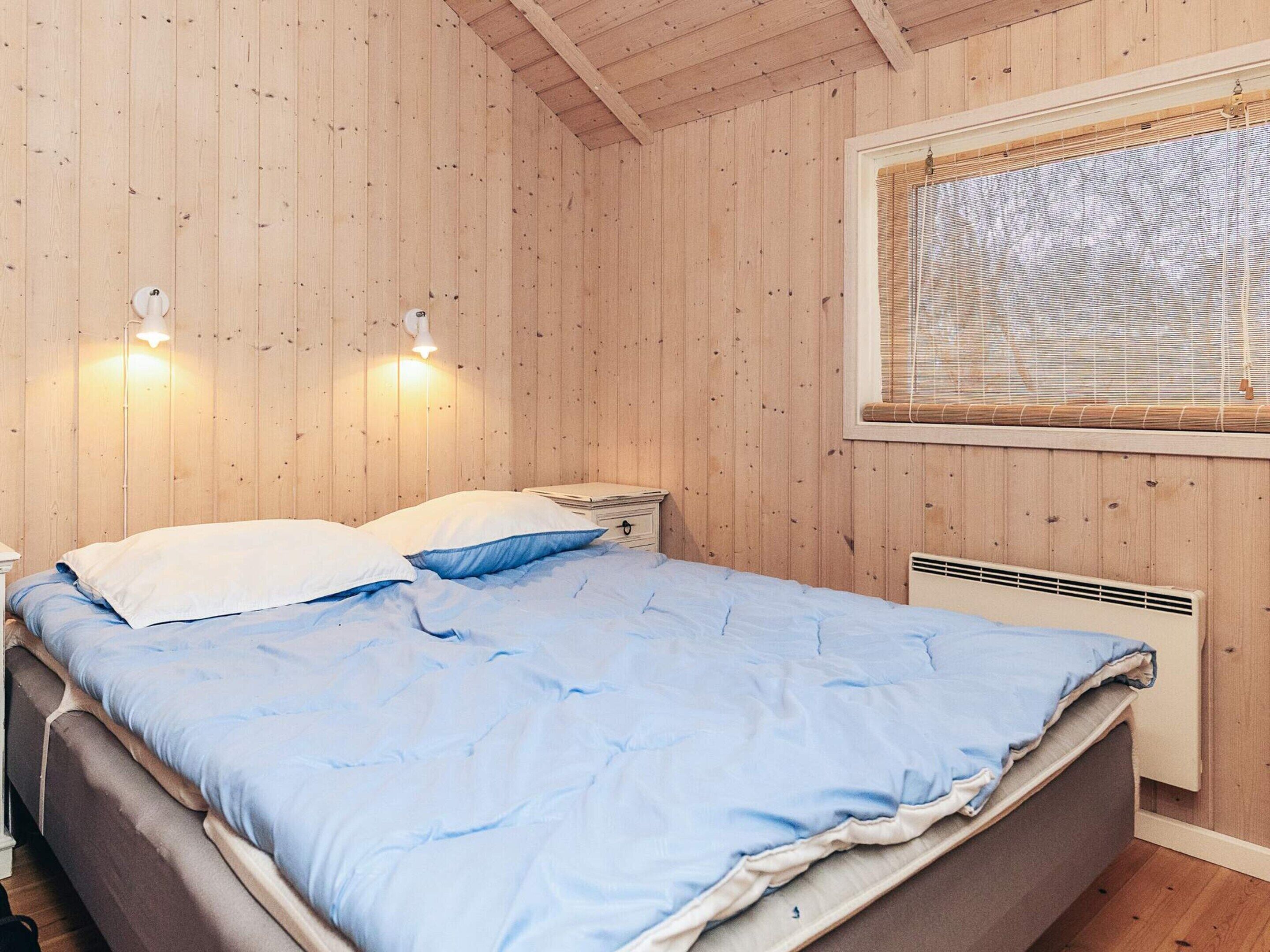 A wooden-paneled room with a bed, two bedside lamps, a window with blinds, and a wall-mounted heater.