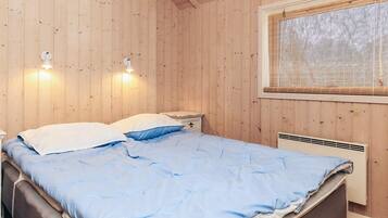 A wooden-paneled room with a bed, two bedside lamps, a window with blinds, and a wall-mounted heater.
