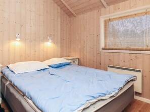 Une pièce aux murs lambrissés, comprenant un lit, deux lampes de chevet, une fenêtre avec stores et un radiateur fixé au mur.