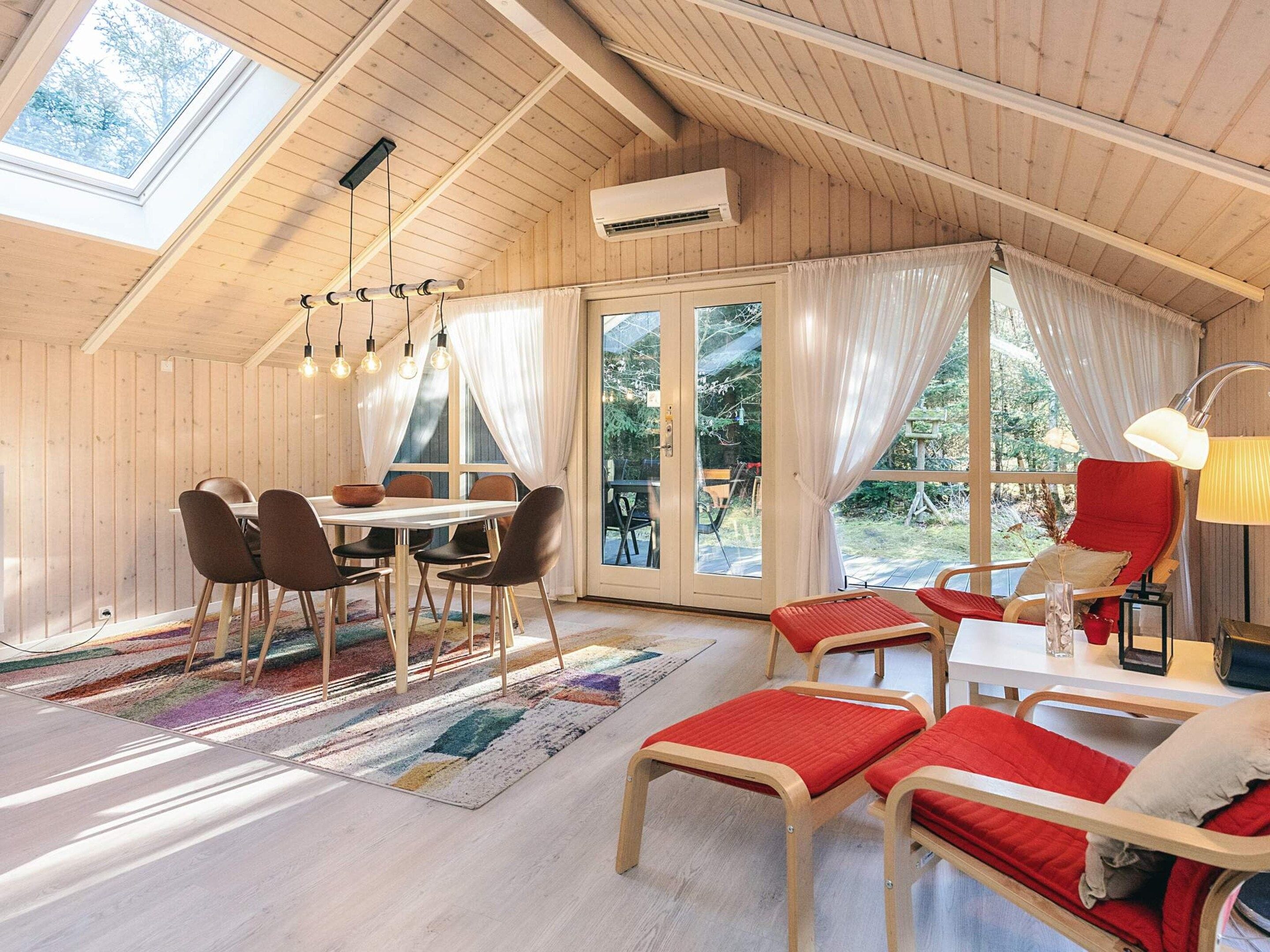 A cozy room with a wooden ceiling, a dining table with chairs, a red chair, a lamp, and a view of the outdoors.
