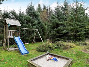A wooden play structure with a slide and swings, surrounded by dense forest.