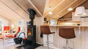 Ein gemütliches Blockhaus-Interieur mit Holzofen, zwei Barhocker, einem Essbereich mit einem roten Stuhl und einer Küche mit einem weißen Kühlschrank.