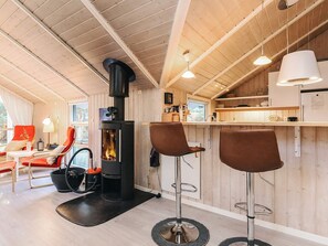 A cozy cabin interior with a wood-burning stove, two bar stools, a dining area with a red chair, and a kitchen with a white refrigerator. - 4 Star Holiday Home in Hals (Hals)
