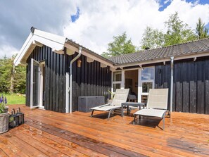 Une terrasse en bois avec deux chaises longues et une petite table, un bâtiment noir avec une porte blanche, et un ciel dégagé.