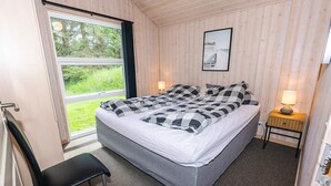 A bedroom with a bed, a chair, a bedside table with a lamp, and a window with a view of greenery.