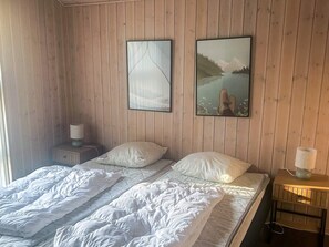 Quarto com parede de madeira, cama com roupa de cama branca, duas mesas de cabeceira com abajures e duas imagens emolduradas.