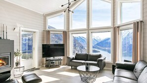 A modern living room with a fireplace, a flat-screen TV, a sofa, a coffee table, and large windows offering mountain views. - Holiday Home in Valldal (Fjord)
