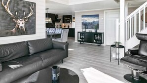 A modern living room with a black leather sofa, a coffee table, and a painting of a deer. There is a dining area with a table and chairs, and a kitchen in the background. - Holiday Home in Valldal (Fjord)