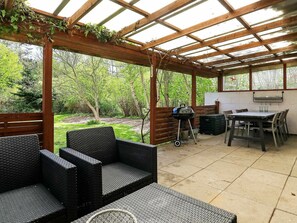 Ulkoterassi, jossa on pergola, rottingista valmistetut huonekalut, grilli ja ruokapöytä tuoleineen.