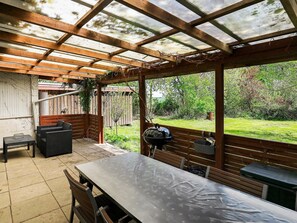 Een overdekt terras met een tafel, stoelen, een barbecue en uitzicht op een tuin.