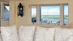 A room with a large bay window overlooking a beach, a wall-mounted clock, and a sofa with floral cushions. - Seaside Charm at Binderup - By Traum Ferienwohnungen (Bjert)