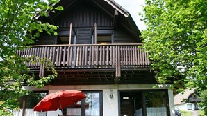 A two-story wooden house with a balcony, a red umbrella, and a table with chairs outside. - Beautiful apartment in villa for 3 people with WIFI, TV and terrace (Korbach)