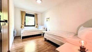 A small, well-lit room with two beds, a window with curtains, a wall-mounted light, and a small table with a light bulb. - Cosy apartment in villa for 4 people with WIFI, A/C, TV, terrace and pets allowed (Bad Wildungen)