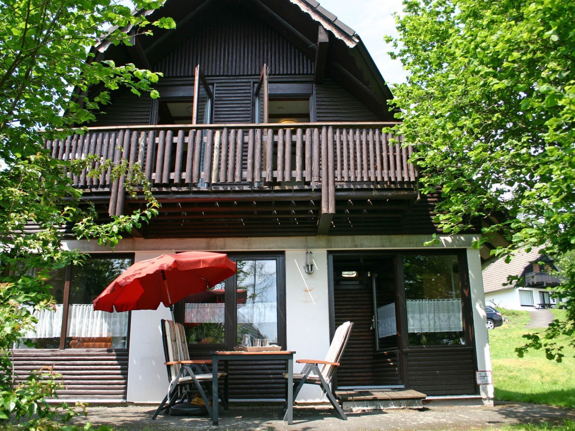 Ein zweistöckiges Holzhaus mit Balkon, rotem Sonnenschirm und Tisch mit Stühlen vor der Fassade.